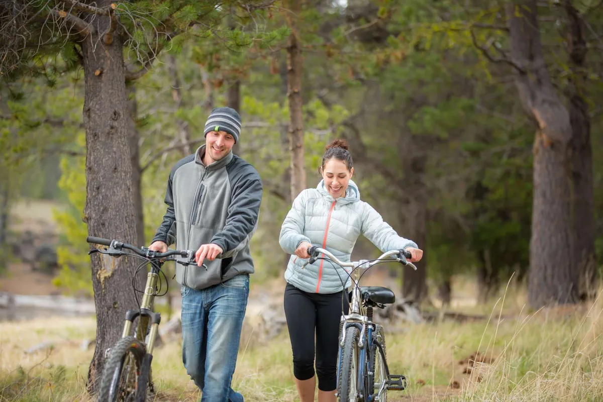 The image shows two people walking their bicycles along a grassy path lined with tall trees in the background. The atmosphere is joyful and relaxed, capturing the spirit of outdoor exploration and easy trail access.