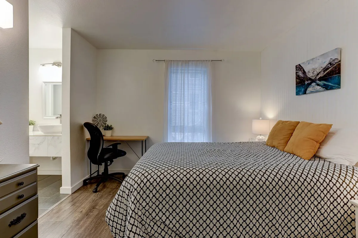This image shows a comfortable bedroom featuring a cozy bed, a dedicated desk set up by the window, and curated artwork on the walls. Natural light streams in, creating a soothing and productive environment ideal for both work and relaxation.