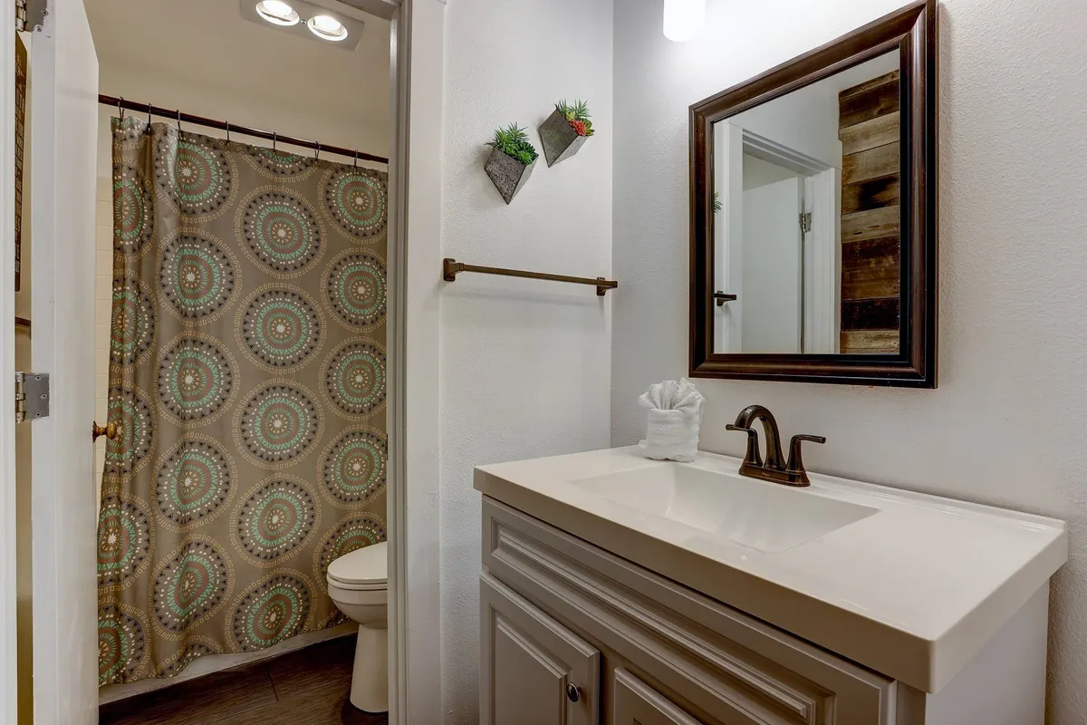 A spacious bathroom attached to a bedroom, featuring a modern vanity with a large mirror, a bright shower curtain, and a clean, inviting ambiance. The atmosphere is peaceful and private, perfect for unwinding and self-care.
