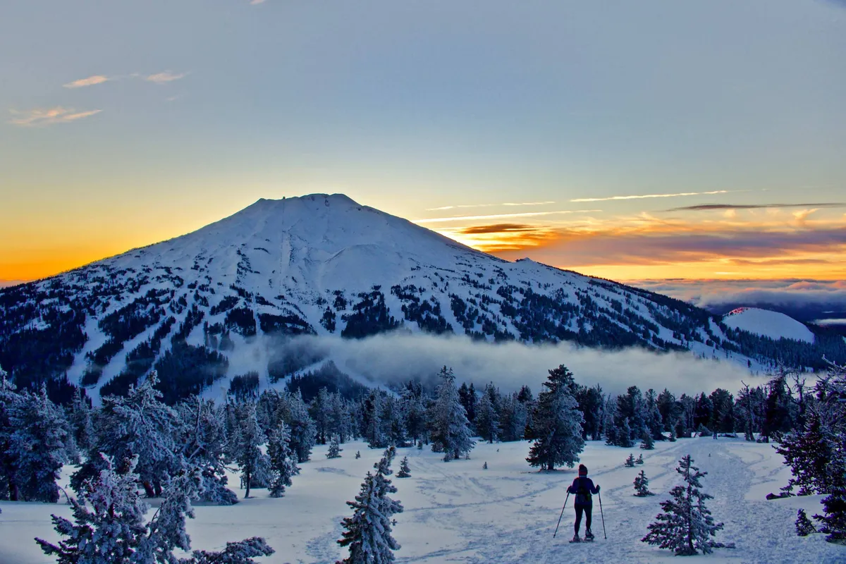 A dynamic winter landscape featuring a skier gliding down a snow-capped mountain at sunset. The vibrant sky casts a golden glow over the snowy slopes, capturing the thrill and serenity of winter sports in the Cascades.