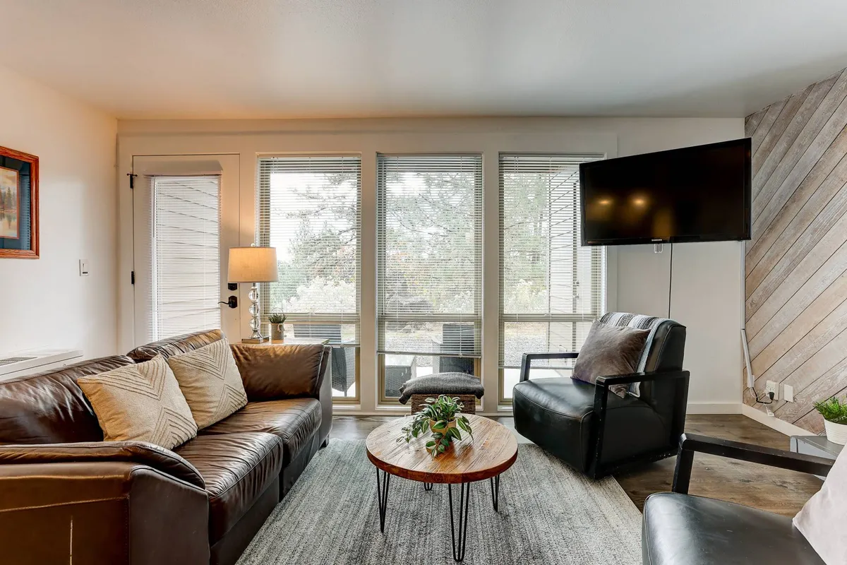 This image features a cozy living area with a leather sofa, coffee table, accent chairs, and large windows that frame scenic woodland views. The space feels inviting and comfortable, ideal for relaxing after outdoor adventures.