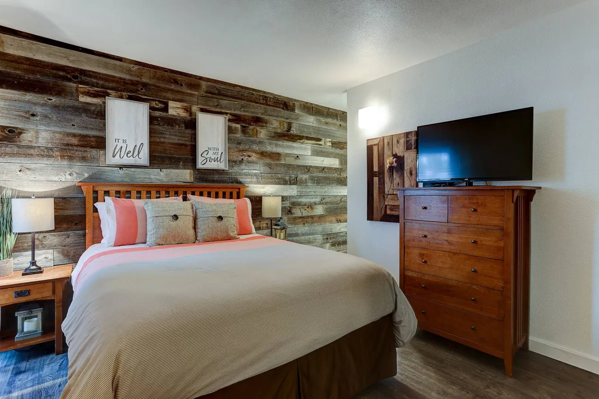 A serene bedroom interior featuring a wooden accent wall, a plush bed with decorative pillows, a nightstand with a glowing lamp, and a wall-mounted television. The atmosphere is calm and inviting, ideal for relaxation and unwinding after a busy day.