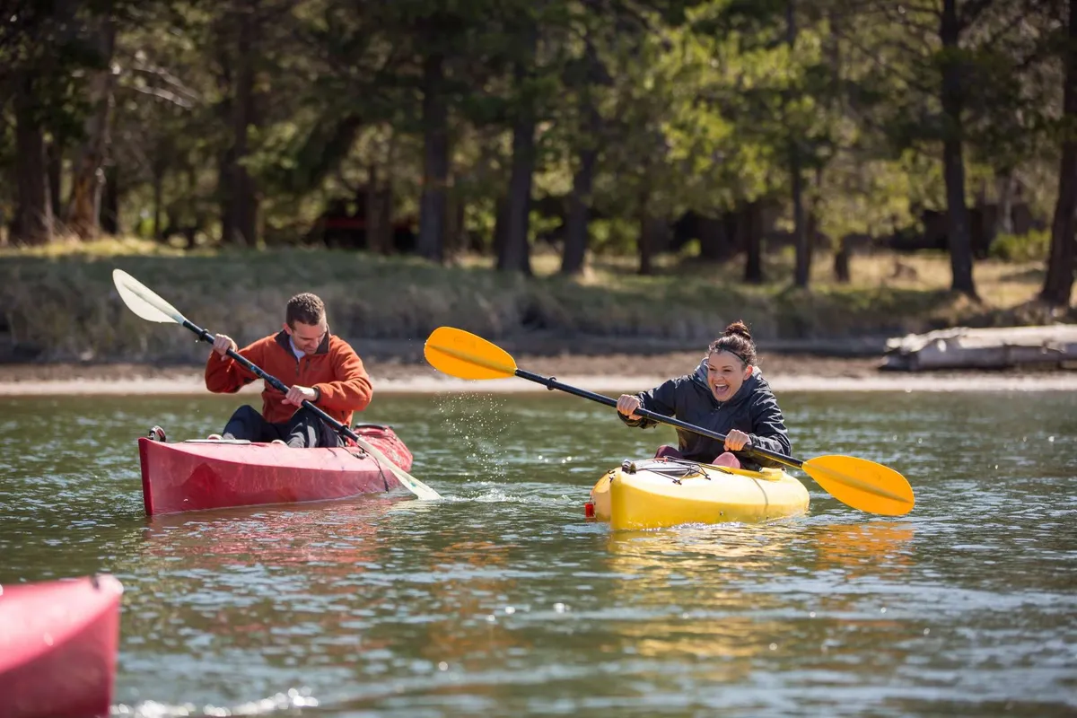 Two brightly colored kayaks cut through the serene waters, their reflections mirrored beneath them. The kayakers are enjoying a peaceful moment surrounded by nature, with the water and lush banks creating an inviting outdoor scene.