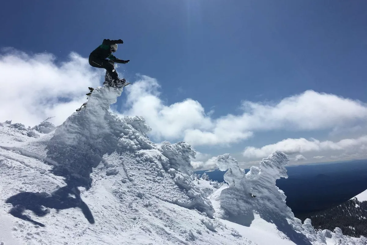 An action shot of a snowboarder mid-jump against the backdrop of a snowy mountain peak and dramatic clouds. The image captures the thrill of outdoor adventure and the natural beauty surrounding Bend.