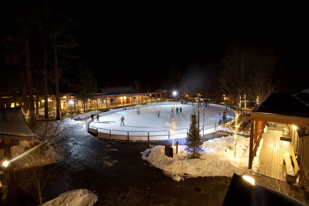 The image features a charming ice skating rink nestled among trees with festive lights and cozy buildings nearby. People are seen skating and socializing, with the joyful ambiance heightened by seasonal decorations, suggesting the magic of winter activities at the resort.