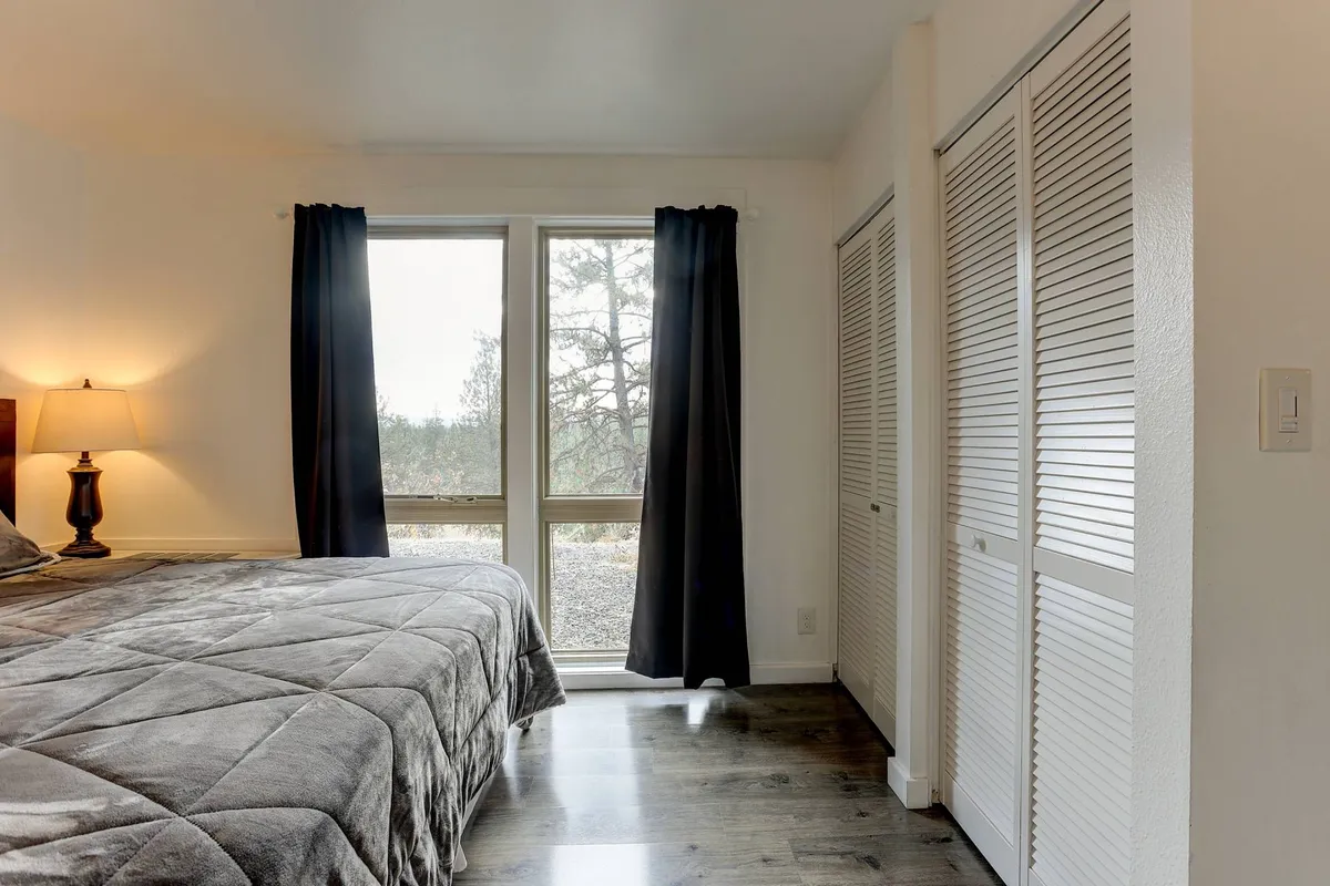 A tranquil bedroom with a gray-quilted bed, a bedside lamp, and expansive windows dressed in curtains. The windows draw in ample natural light and hint at scenic outdoor views, suggesting a seamless link to nature from within the living space.