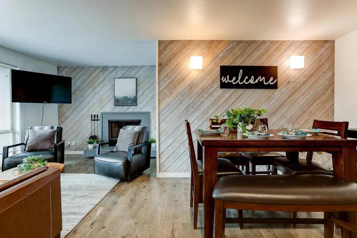 A warm and welcoming interior scene centered around a dining table set for a meal, with a fireplace and a comfortable seating area nearby. A 'welcome' sign and tasteful décor create a cozy, inviting atmosphere ideal for gathering and storytelling.