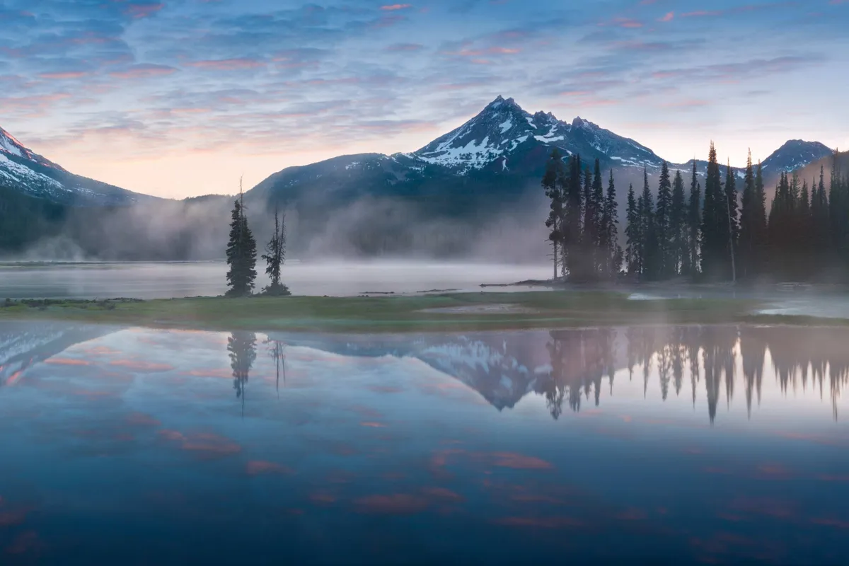 A tranquil view of mist rising from a mountain lake at sunrise, framed by evergreen trees and distant peaks. Trails wind around the water’s edge, inviting exploration, fishing, and scenic relaxation amidst nature.