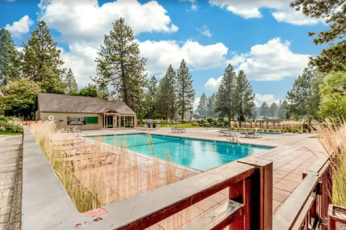 This image showcases a vibrant outdoor swimming pool surrounded by lush greenery and a modern community building in the background. The scene features sunbathers lounging by the pool and families enjoying the water, encapsulating the resort’s lively, social summer atmosphere.