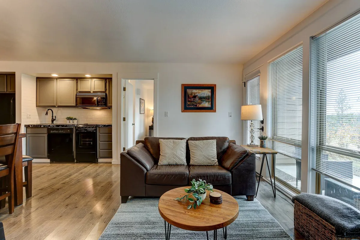 This image features an open-concept space including a leather sofa, a dining table, and a clear view into the kitchen area. Expansive windows line the wall, flooding the area with natural light and offering glimpses of the surrounding woodland, while tasteful artwork adds a homey touch.