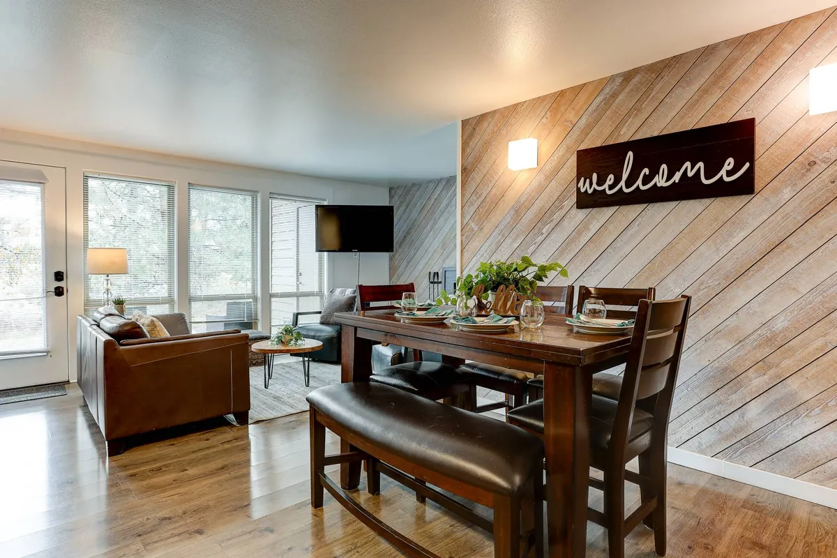 The image showcases a bright, inviting living and dining area with a cozy sofa, a welcoming sign, a spacious dining table perfect for shared meals, and large windows that let in abundant natural light. The setting highlights comfort, togetherness, and relaxation, all with a view of the outdoors just beyond the glass.
